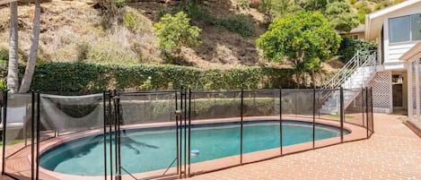 A swimming pool with a safety fence, surrounded by a brick patio, and a hillside with vegetation in the background.