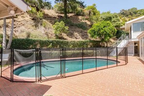 A swimming pool with a safety fence, surrounded by a brick patio, and a hillside with vegetation in the background. - 90210 NEST: Private Pool Retreat in Beverly Hills (Beverly Hills)