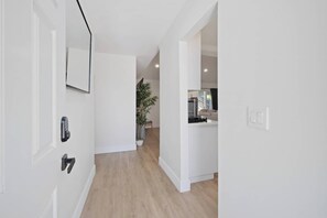 A hallway with a door, a potted plant, and a glimpse of a living area with a sofa and a window. - Spacious 4 Bedroom House W/ Amazing Backyard (Chula Vista)
