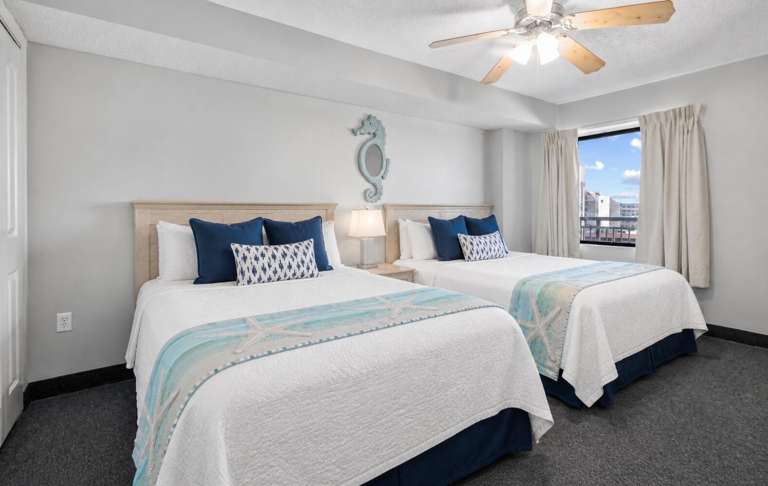 A hotel room with two beds, a ceiling fan, a window with curtains, and a seashore view.