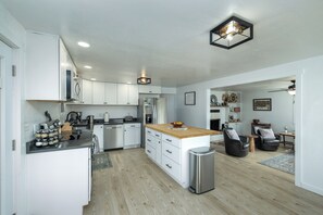 A modern kitchen with white cabinets, a wooden countertop, and a small island. There is a refrigerator, a sink, and a coffee maker. The kitchen connects to a dining area with a wooden floor and a small table.