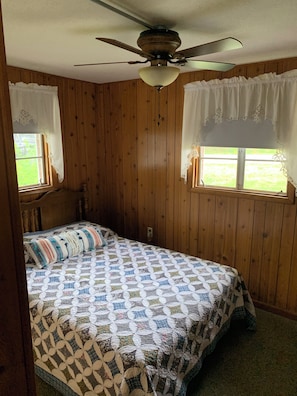 Quarto com paredes de madeira, cama com colcha estampada, ventoinha no teto e duas janelas com cortinas de renda.