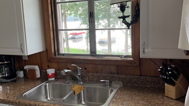 Una cocina con encimera de granito, pileta de acero inoxidable, paneles de madera y una ventana con vista a árboles y un vehículo rojo en el exterior.