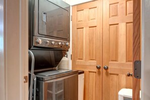 A compact laundry room with a washer and dryer, wooden double doors, and a small trash bin. - KBM Resorts | Town Pointe | Spacious 1 level, Walk to Main and Town Lift, Private Hot Tub, Parking, Elevator Access, A/C! TPC-102c (Park City)
