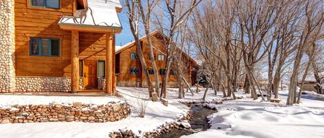 A log cabin with a stone foundation, a wooden porch, and a stream running through the snow.