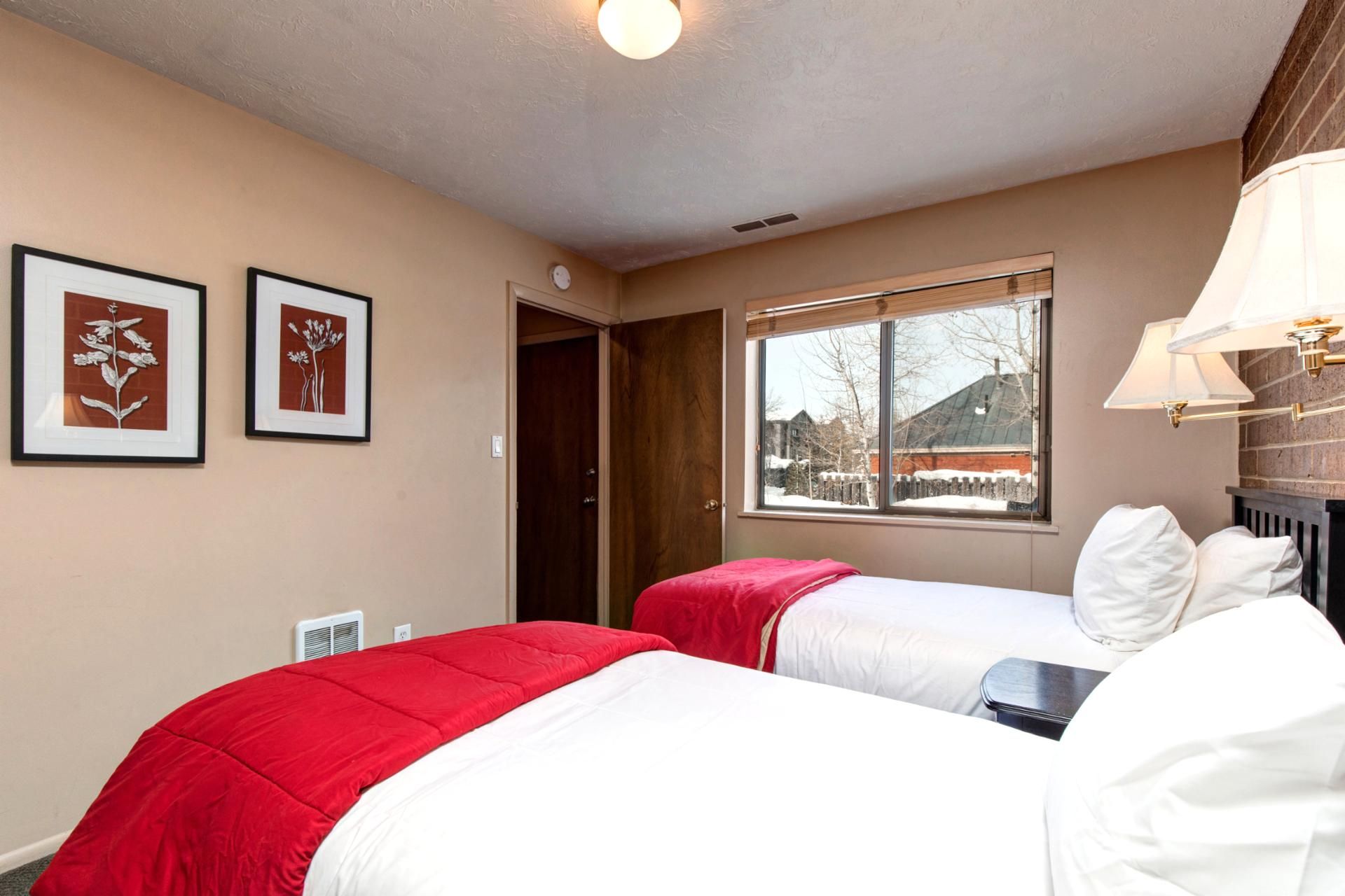 A hotel room with two beds, a window with a view of buildings and trees, and framed artwork on the wall.