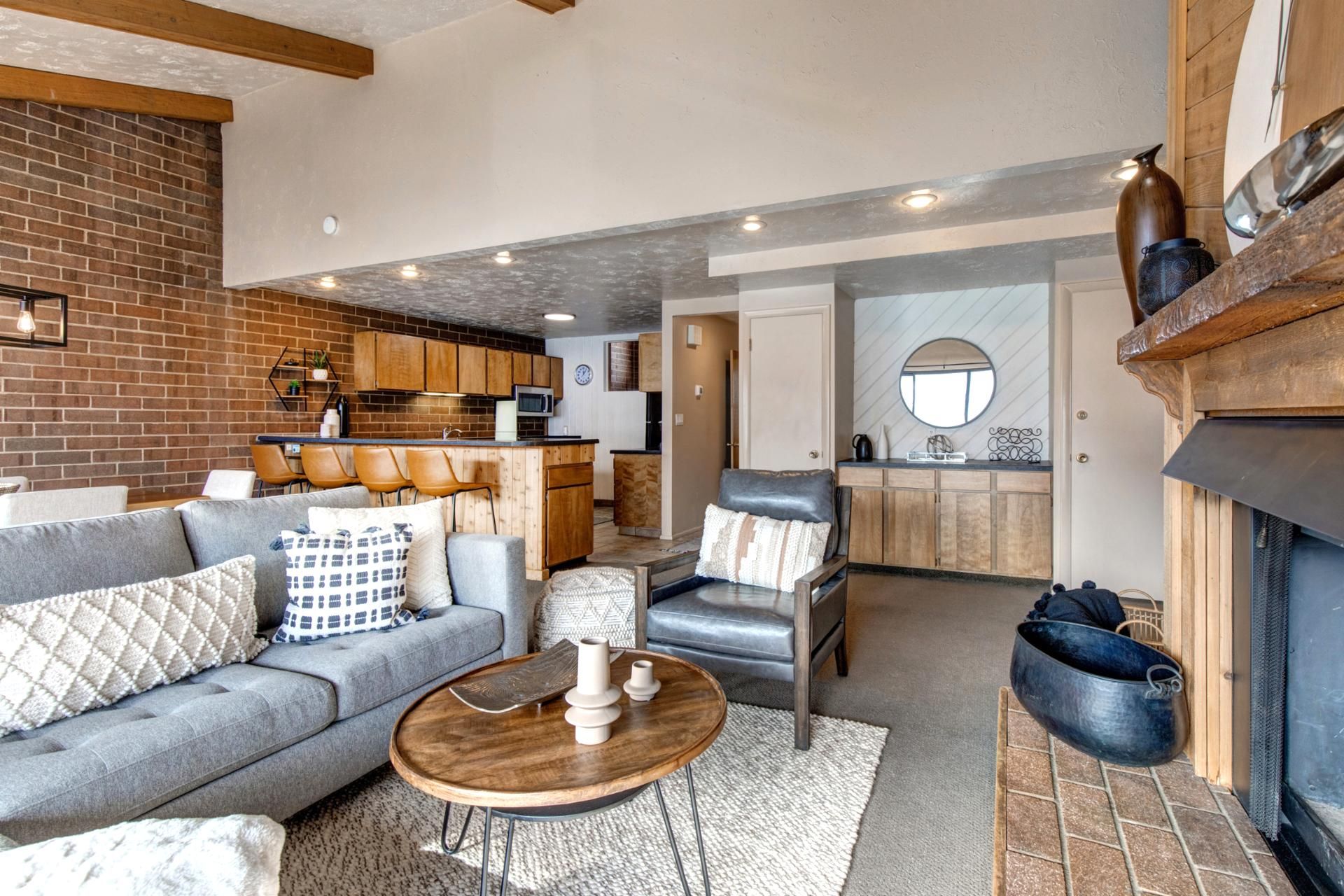 A living room with a sofa, armchairs, a coffee table, and a fireplace. There is a kitchen in the background with wooden cabinets and a microwave.