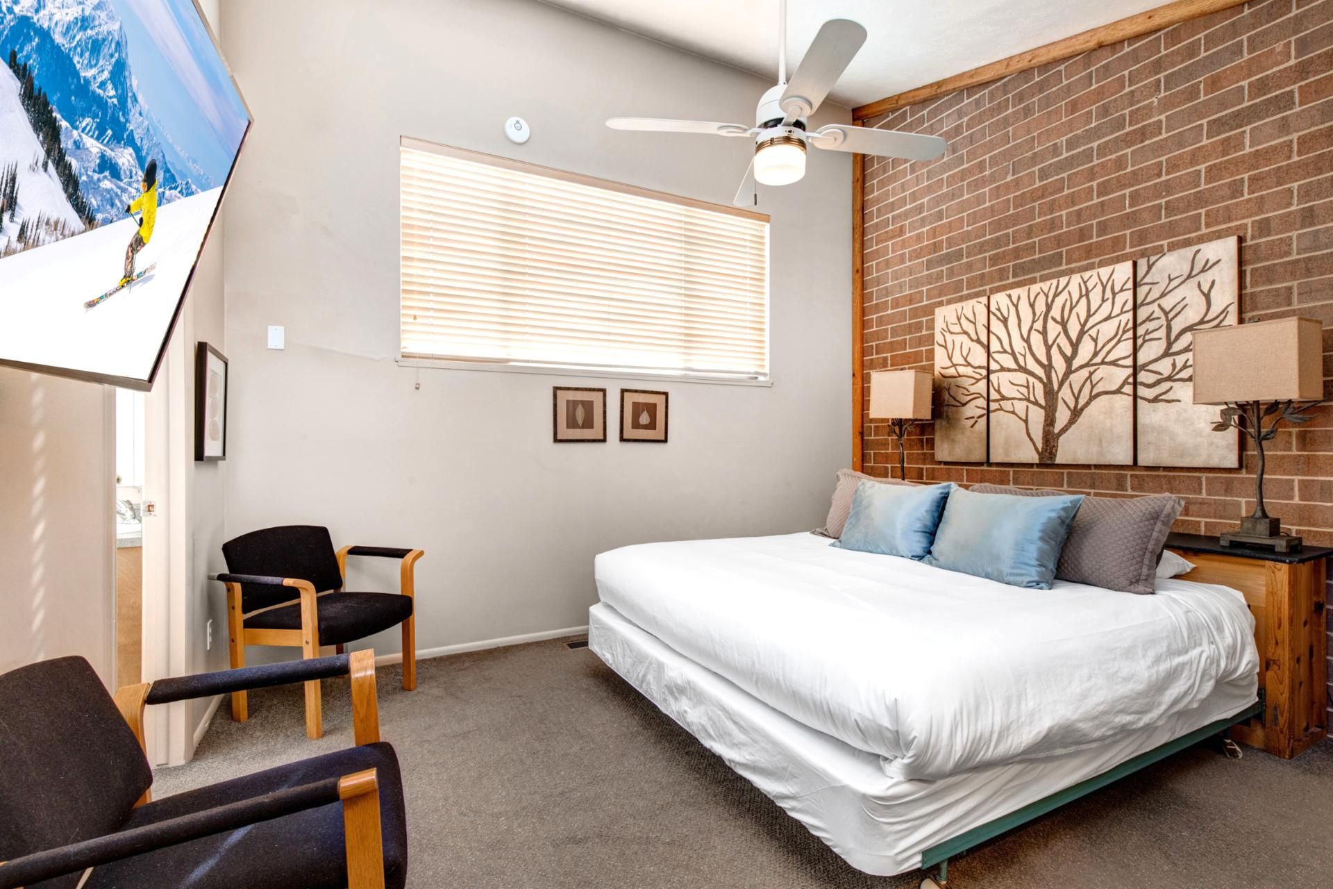 A bedroom with a bed, a ceiling fan, a window with blinds, a chair, and a wall with a tree painting.