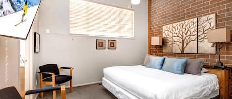 A bedroom with a bed, a ceiling fan, a window with blinds, a chair, and a wall with a tree painting.
