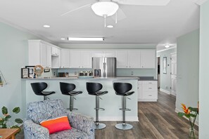 A modern kitchen with white cabinets, a stainless steel refrigerator, and a bar area with black stools. - OW 1303-Luna Sol: Experience coastal charm in Oak Island, a spacious 2BR condo with ocean views! (Oak Island)