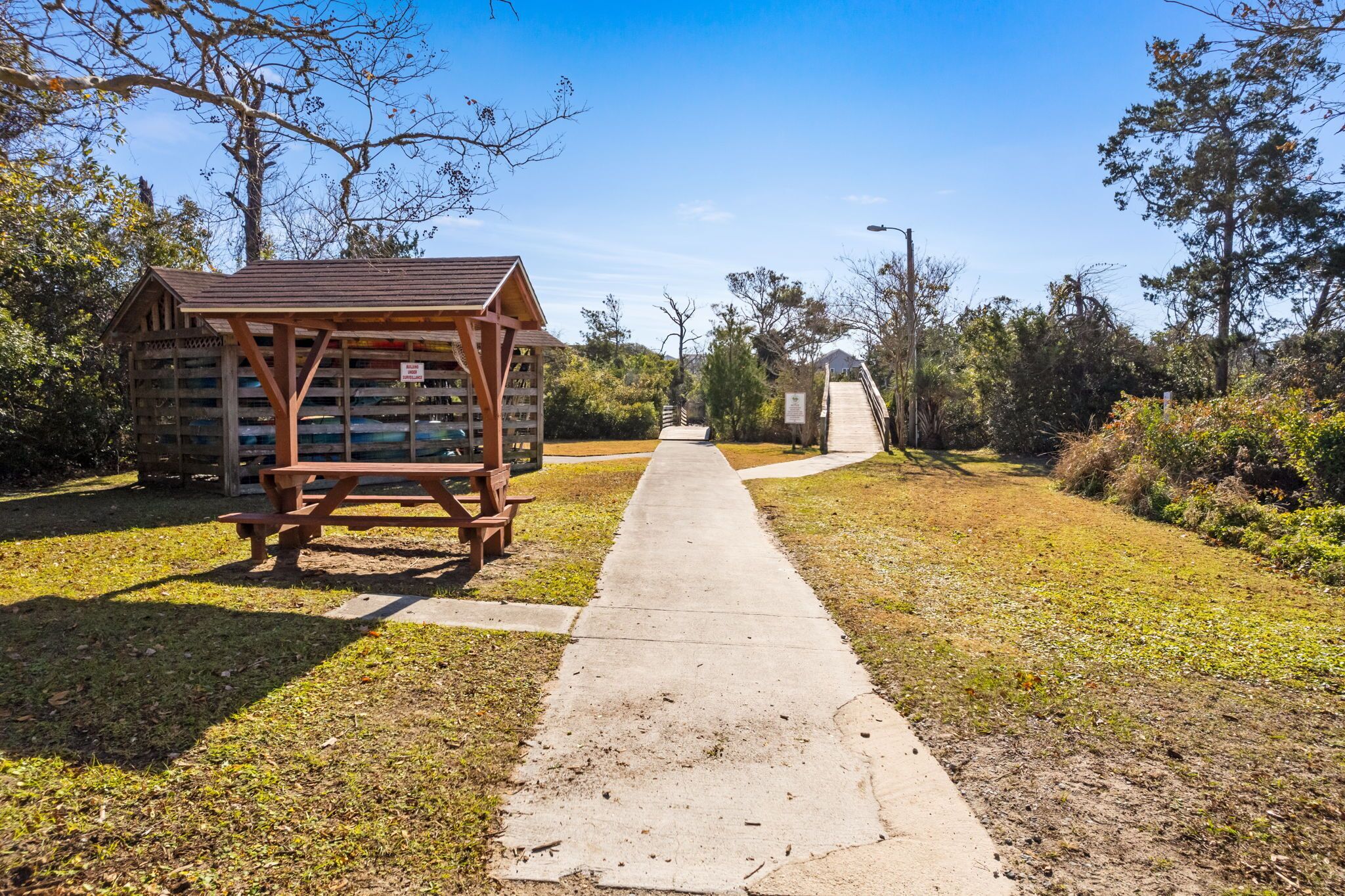 A picnic area with a wooden structure and benches, a concrete path, and a bridge in the background.