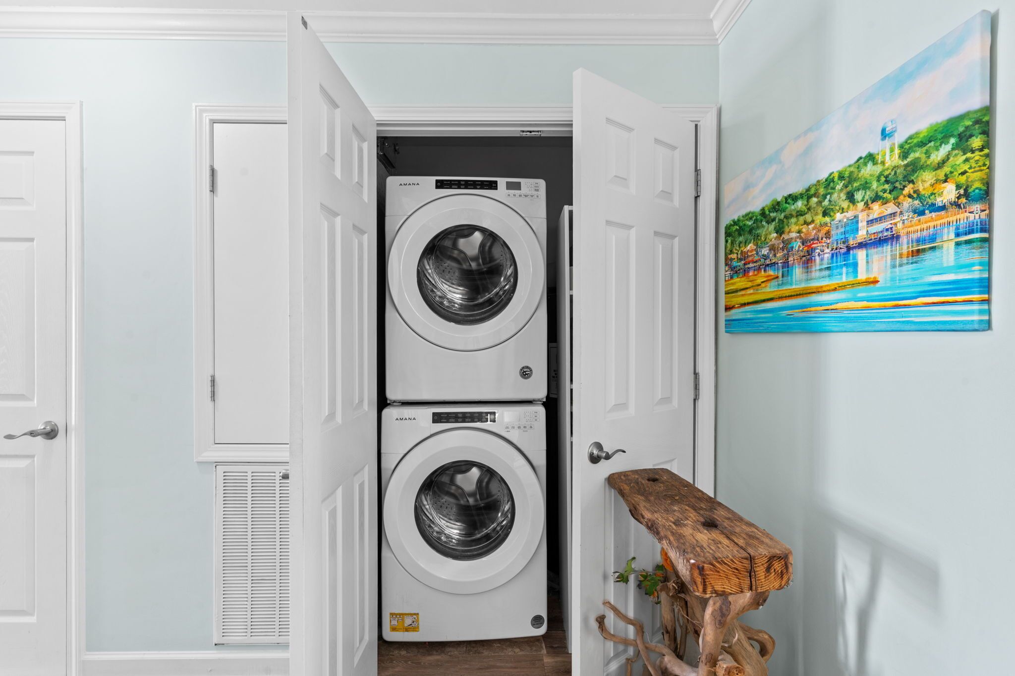 A laundry room with two stacked front-loading washing machines, a wooden bench, and a painting on the wall.