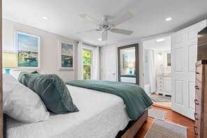 Ein Schlafzimmer mit einem Bett, einem Deckenventilator, einem Fenster mit einem Gemälde und einer Tür zu einem anderen Raum.