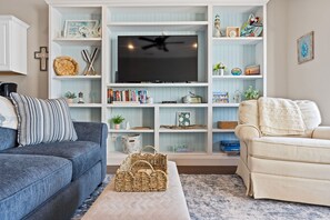 Ein Wohnzimmer mit Fernseher, zwei Sofas, Regalen mit Büchern und Dekoration sowie einem Deckenventilator.