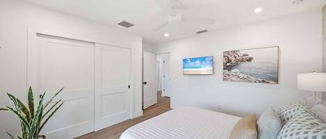 Un dormitorio con una cama, ventilador de techo, televisor montado en la pared y un cuadro enmarcado.