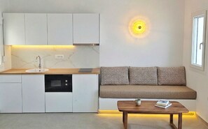 A compact kitchen with a built-in dishwasher, a sofa with patterned cushions, and a coffee table with a book. - Martynou View  (Santorini)