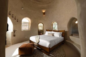 A bedroom with a wooden bed, a small ottoman, and arched windows. - Sandora Domes - Architectural Retreat Near National Park (Joshua Tree)