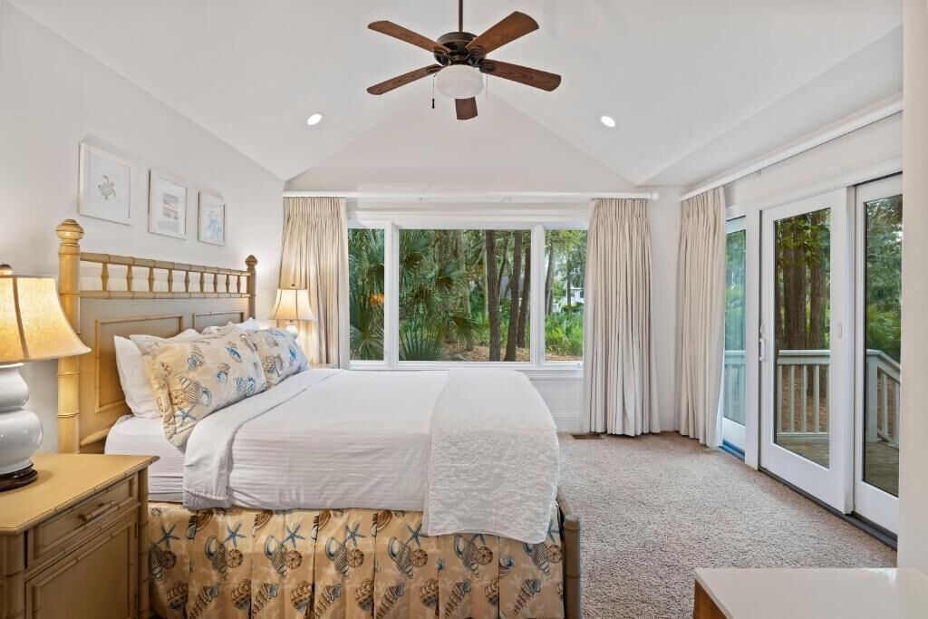 A bedroom with a bed, a ceiling fan, a window with curtains, and a view of greenery outside.