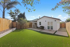 A residential property with a fenced backyard, a patio with outdoor furniture, and a white building with a red roof. - Cozy Luxury Retreat: Near the Beach & Garden Views (San Diego)