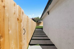 A narrow pathway flanked by wooden fences and a white wall, leading to a grassy area with a tree. - Cozy Luxury Retreat: Near the Beach & Garden Views (San Diego)