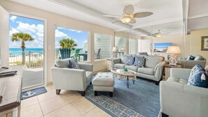 A spacious living room with a large window overlooking a beach, a ceiling fan, and a television. - Gulfside Gem 144-Gulf Views-Sleeps 6 (Panama City Beach)