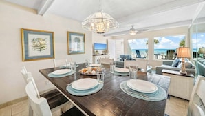 A dining area with a wooden table set for four, framed botanical prints on the wall, and a view of the ocean through large windows. - Gulfside Gem 144-Gulf Views-Sleeps 6 (Panama City Beach)