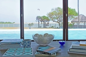 Une table avec un saladier de fruits, un bol bleu et des livres, donnant sur la mer et les palmiers à travers de grandes fenêtres.