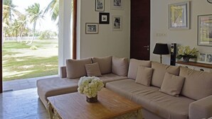 Un salon avec un canapé beige, une table basse en bois ornée d’un vase de fleurs et une vue sur des palmiers, visible à travers une porte vitrée.