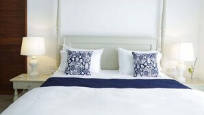 Un lit bien fait, avec des oreillers blancs et bleu marine à motifs, une tête de lit blanche et deux lampes de chevet posées sur une table de chevet blanche.