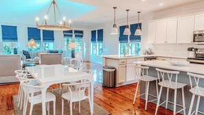 A modern kitchen with white cabinetry, a central island with bar stools, and a dining area with a white table and chairs. - 🦩Book Me Baby! Cottage By The Sea at Sandestin Resort~Golf Cart🦩 (Miramar Beach)
