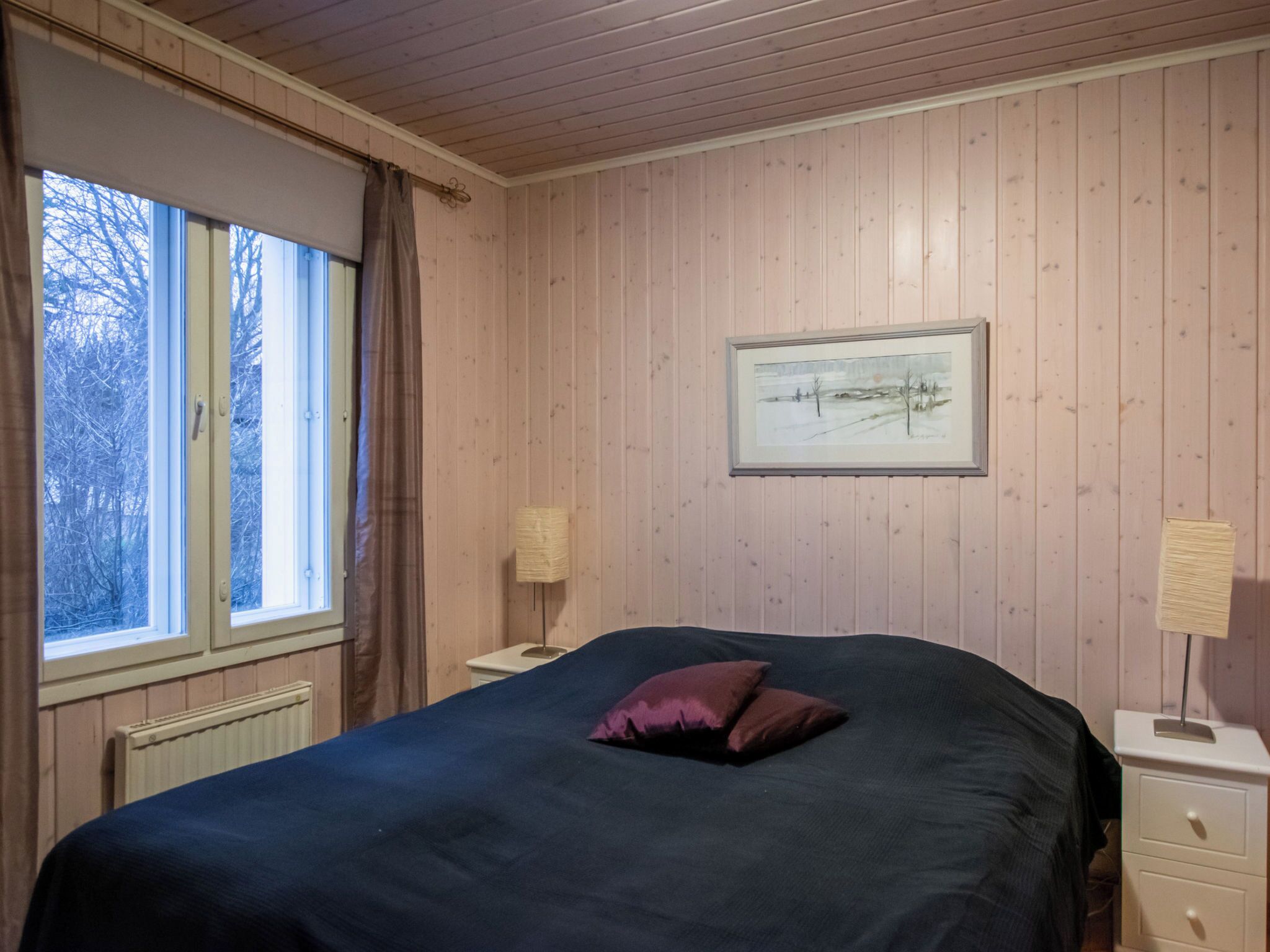 Ein Schlafzimmer mit Bett, Fenster, Heizkörper, Nachttisch, Lampe und einem gerahmten Bild an der Wand.