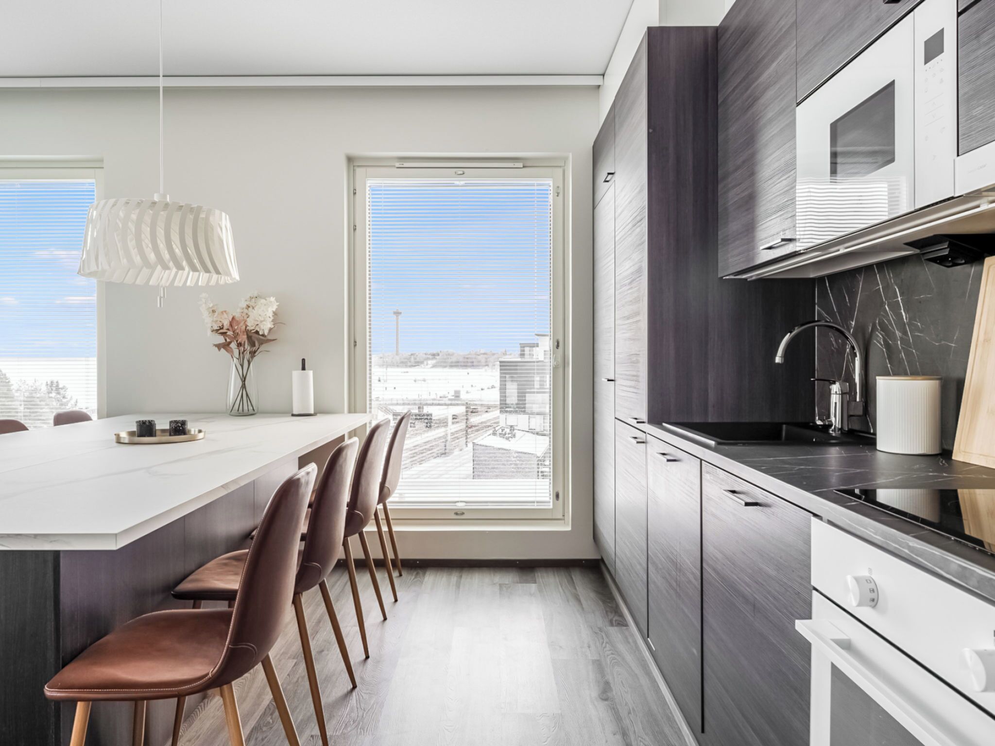A modern kitchen with a large island, dark cabinetry, and a view through a large window.