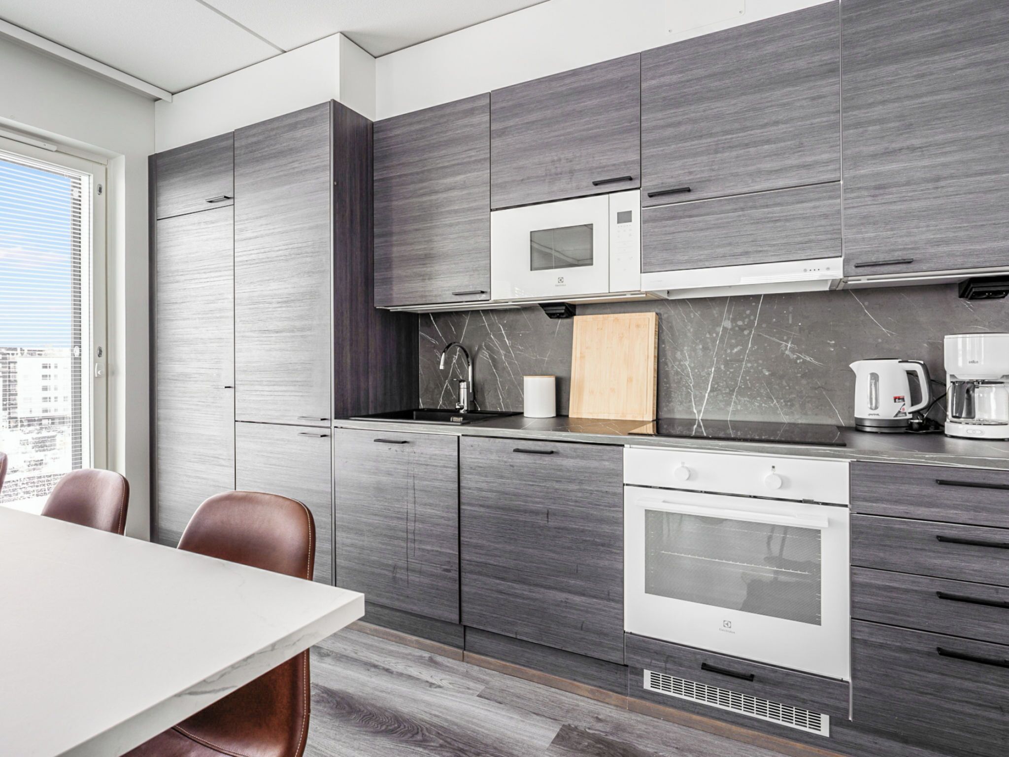 A modern kitchen with grey cabinetry, a built-in microwave, a coffee maker, and a window with a city view.
