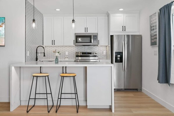Una cocina moderna con armarios blancos, un refrigerador de acero inoxidable, un microondas y dos taburetes.