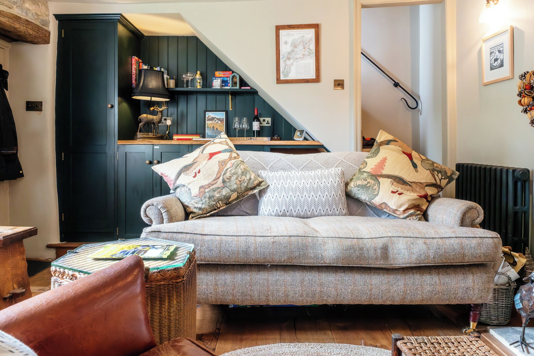 A cozy living room with a sofa, a wicker chair, a small table, and a kitchen area with a staircase.