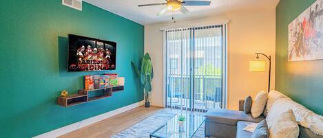 A living room with teal walls, a ceiling fan, a flat-screen TV, a sofa, a coffee table, and a large window with blinds.