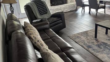 Un salon avec un canapé en cuir marron, une cheminée en pierre, un coin repas avec des chaises et un ventilateur de plafond.
