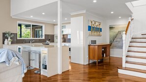 A modern kitchen with a central island, a dining area with bar stools, a staircase, and a view of the outdoors. - Copa Panorama House Stay (Copacabana)