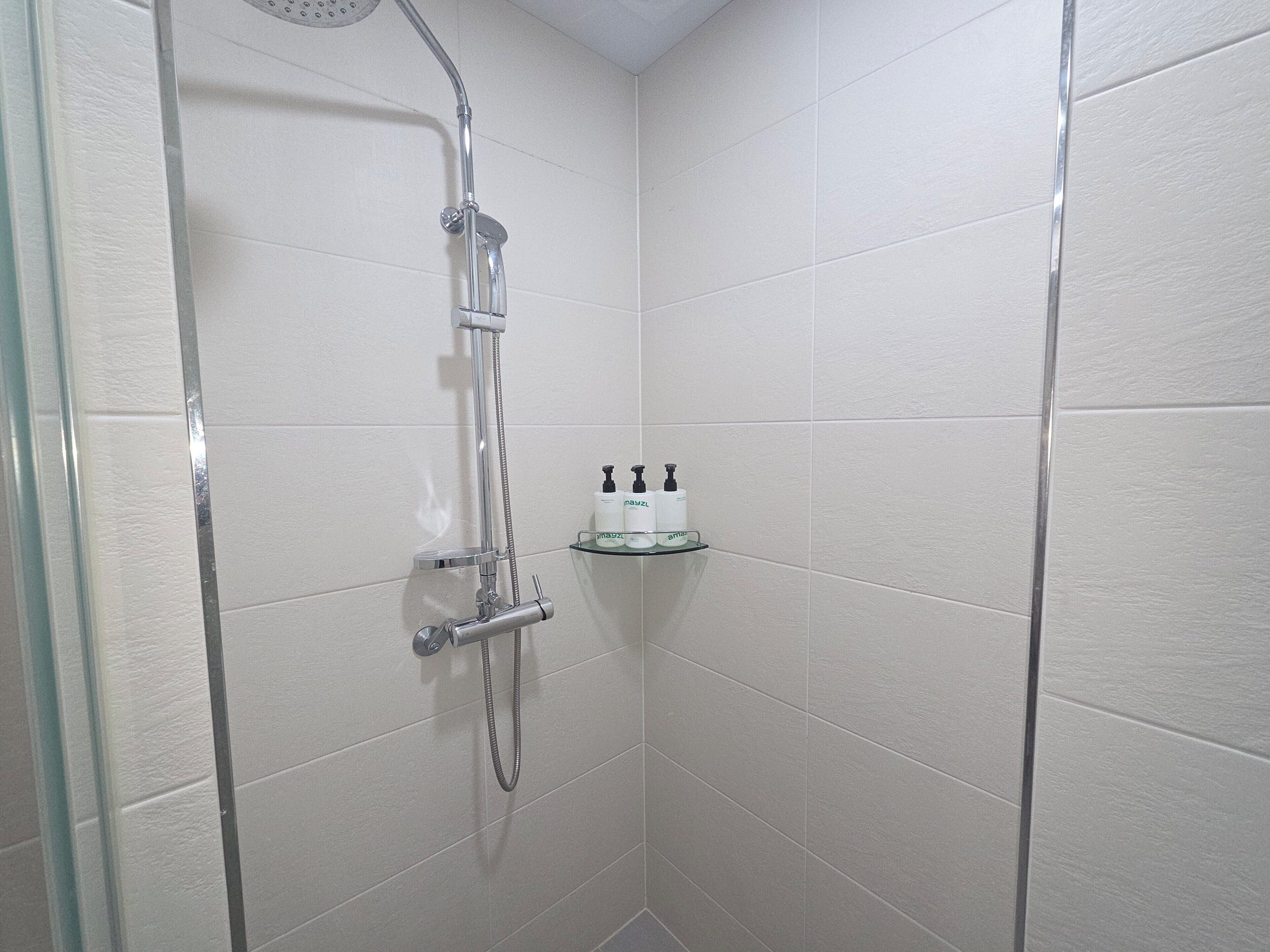 A shower with a handheld showerhead and a wall-mounted faucet, a glass shelf with toiletries, and white tiled walls.
