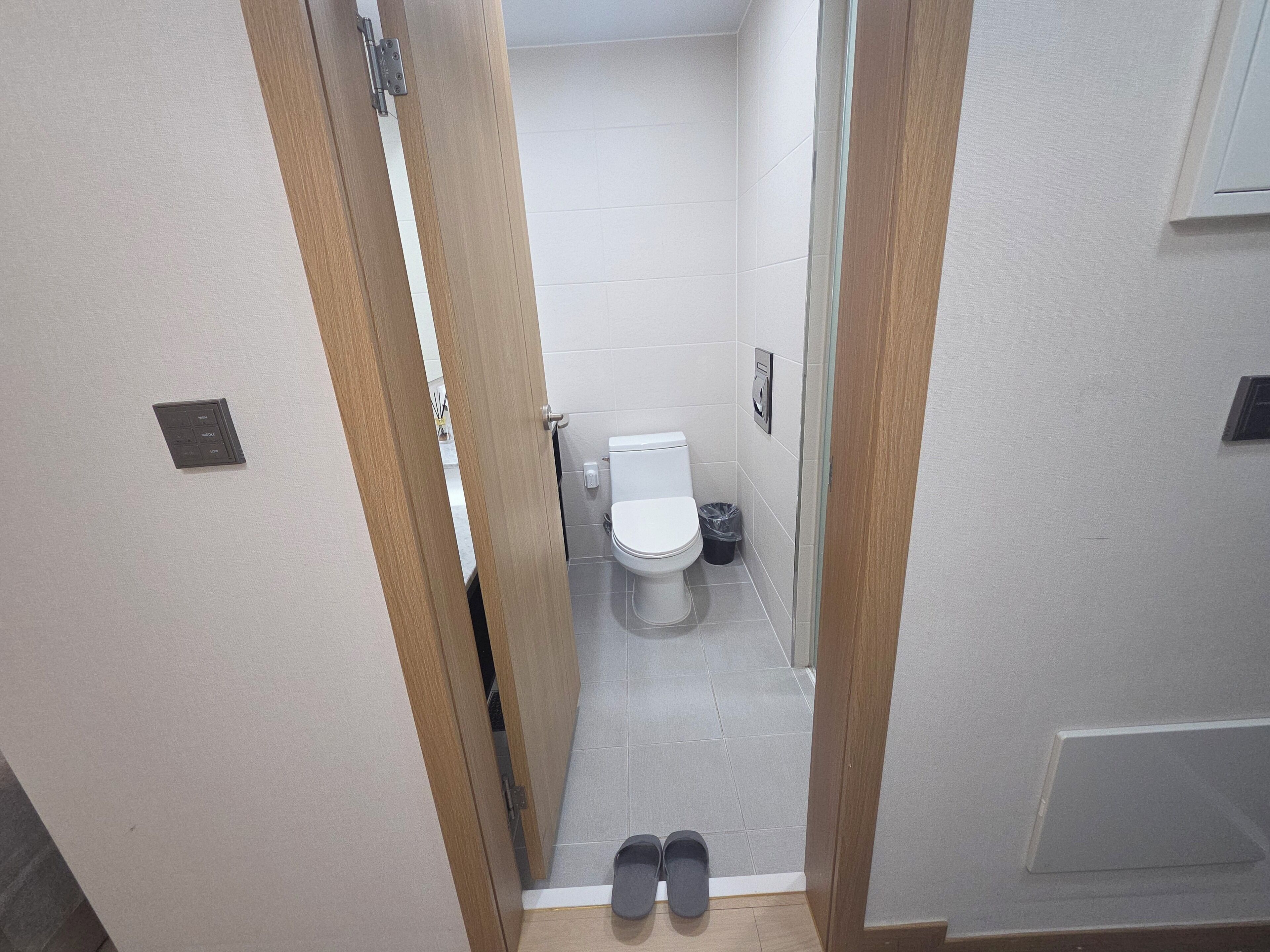 A bathroom with a toilet, a trash bin, and a pair of shoes at the entrance.