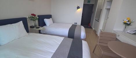 Habitación de hotel con dos camas, una mesita, una silla, un florero con flores y un cuadro en la pared.
