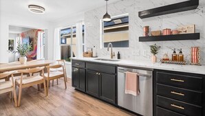 A modern kitchen with a marble backsplash, black cabinets, a stainless steel dishwasher, and a dining area with wooden chairs and a table. - Modern 3BR Home w/ Cowboy Pool + 3-in-1 Game Table (Round Rock)