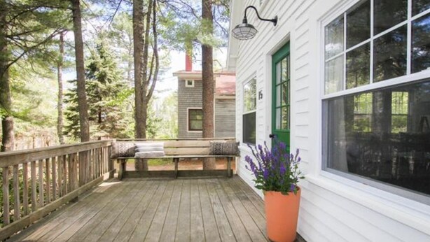 Une terrasse en bois avec un banc, un pot de fleurs et un bâtiment aux murs recouverts de bardage blanc et aux fenêtres vertes.