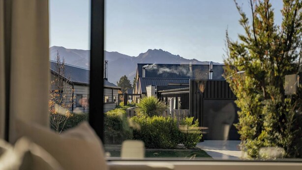 Vista attraverso una finestra di un edificio moderno con montagne sullo sfondo, un giardino con diverse piante e cielo sereno.