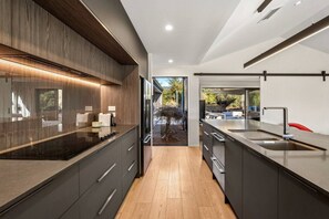 A modern kitchen with dark cabinetry, a stainless steel island, and a view of the outdoor area. - Whare Toi | Designer Stay with Mountain Views (Wānaka)