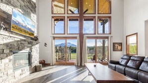 A spacious living room with a stone fireplace, large windows, a leather sofa, and a wooden dining table. - 85-Acre Luxury Lodge | Theater, Hot Tub, Game Room (Island Park)