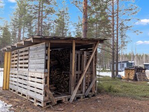 A rustic wooden structure with a slanted roof, a stack of firewood inside, and a clear view of the surrounding forest.