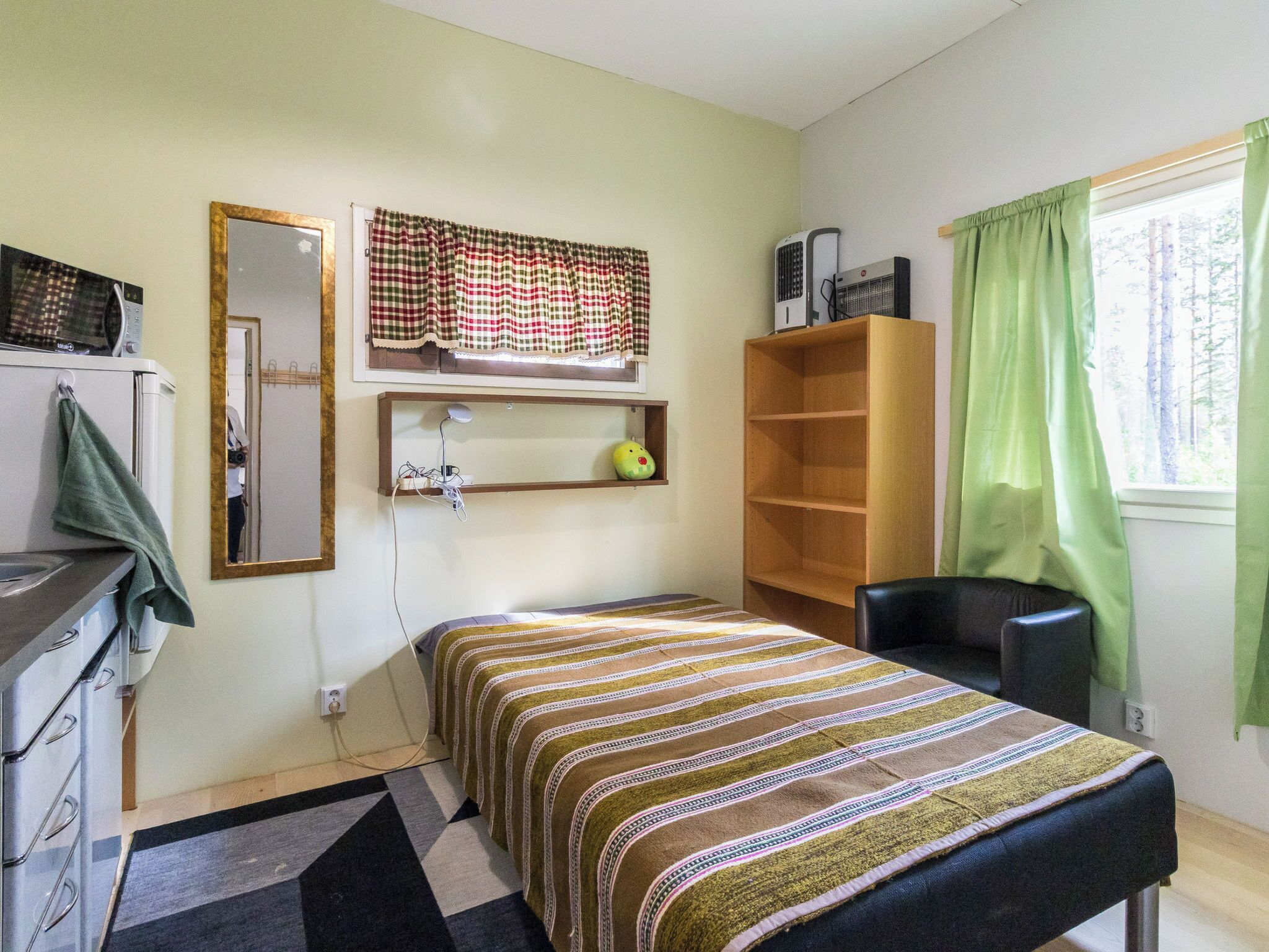 A small, single-bed room with a wooden shelf, a mirror, and a window with curtains.