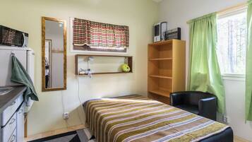 A small, single-bed room with a wooden shelf, a mirror, and a window with curtains.