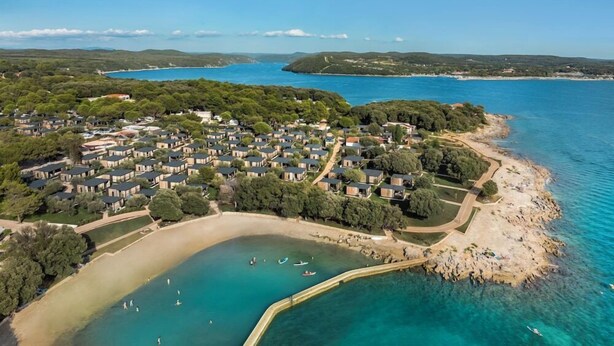 Vista aérea de un complejo turístico costero con varias cabañas, una playa de arena y aguas azules cristalinas.